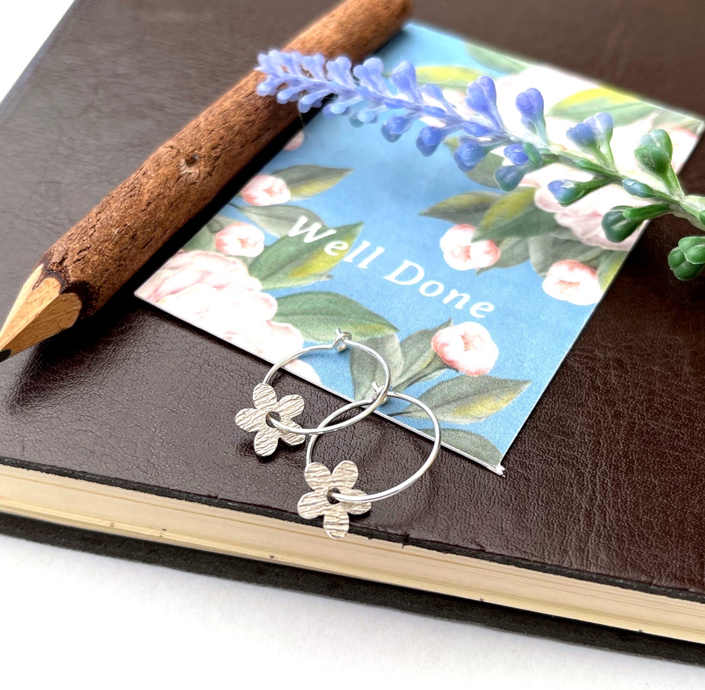 Silver hoop earrings with textured flower charms resting on a blue floral card, styled on a dark brown leather journal with a wooden pencil and small flowers.