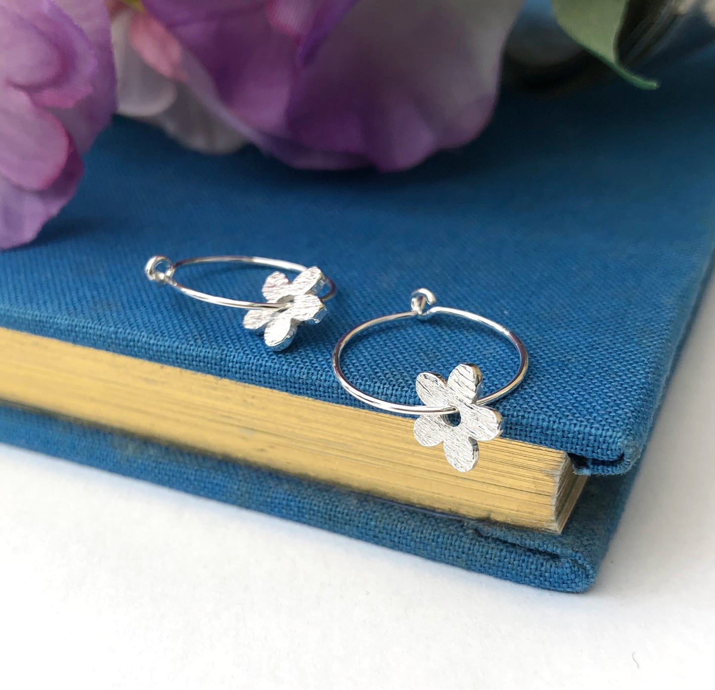 A pair of silver hoop earrings, each adorned with a textured silver flower charm, displayed on a blue book with a wooden edge and purple flowers in the background.