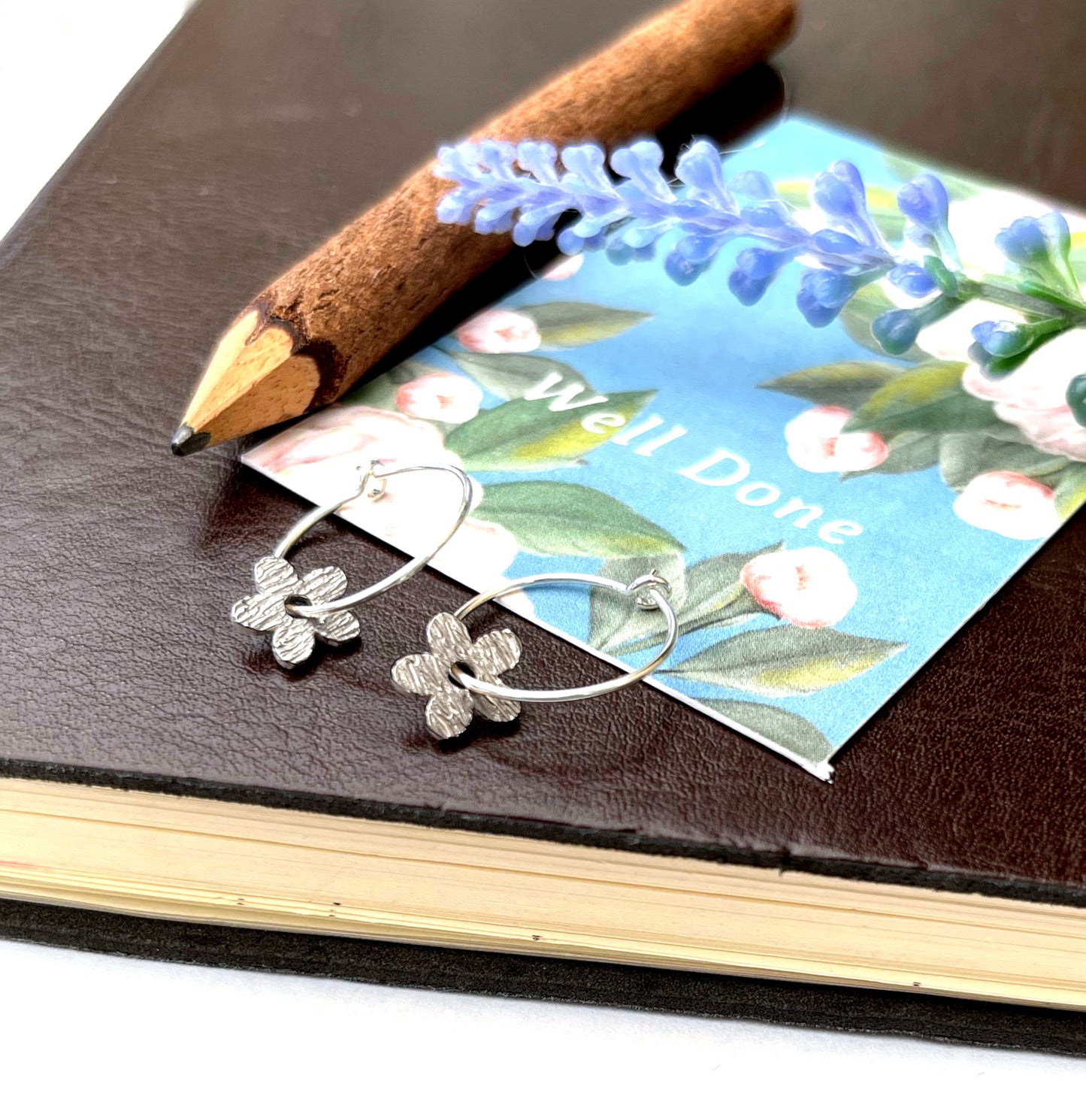 A pair of silver hoop earrings with textured flower charms resting on a blue floral card, placed on a dark brown leather notebook with a wooden pencil.