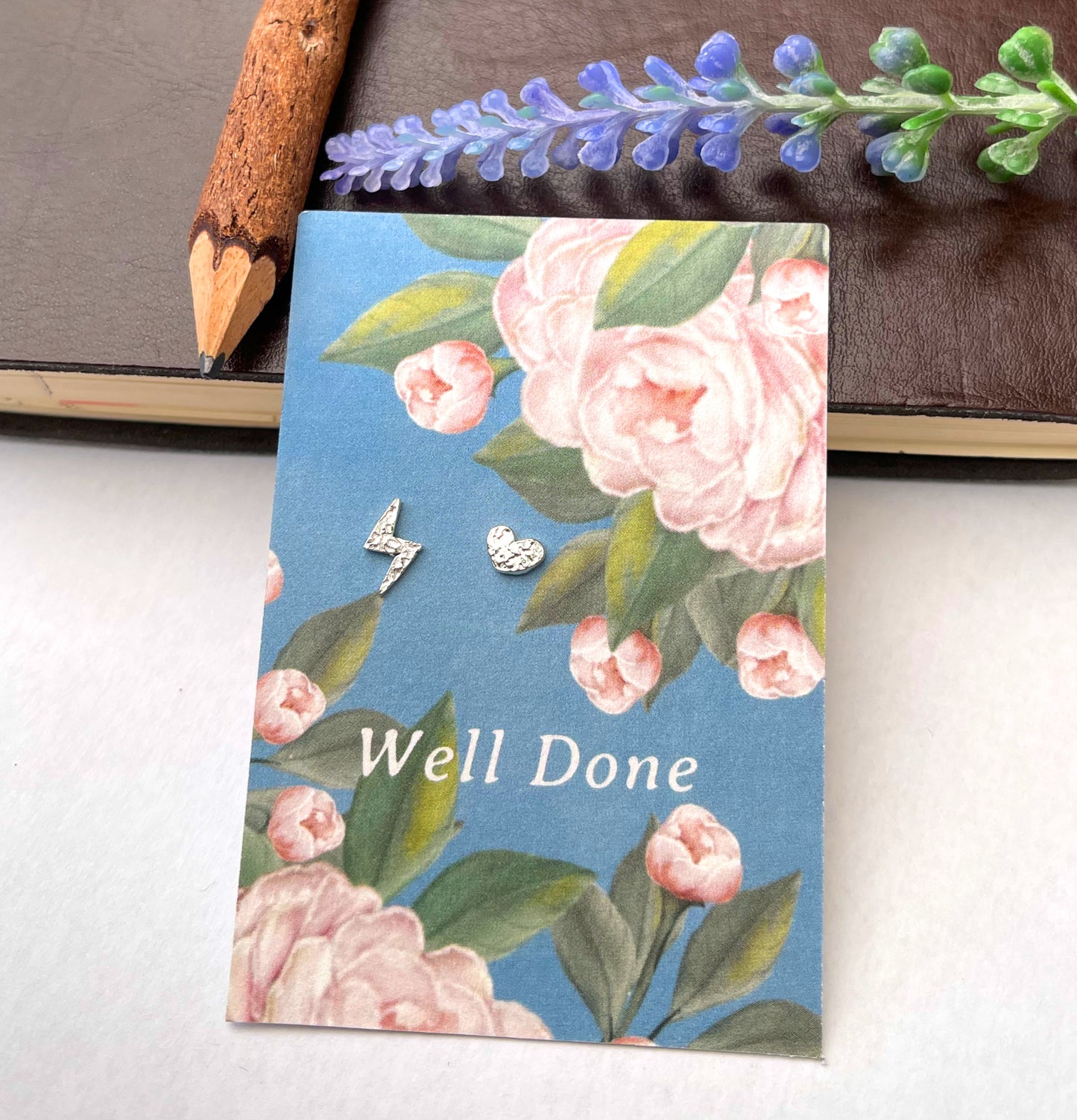 A floral card with the words 'Well Done' in white text, placed on a light surface. The card has a blue background with pink flowers and green leaves, and two small silver stud earrings are resting on it.