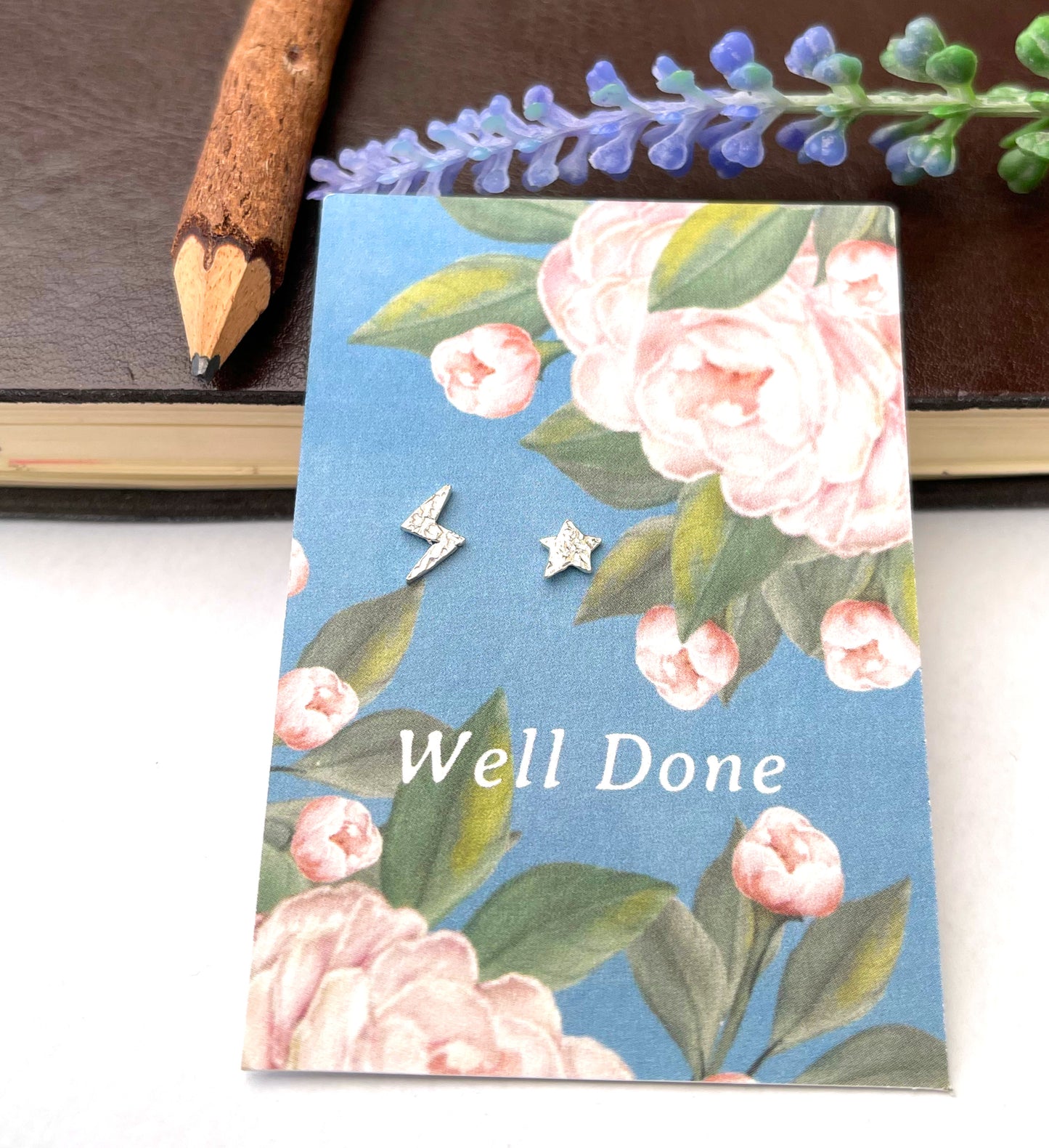 A pair of small silver star and lightning bolt stud earrings displayed on a floral blue card that reads 'Well Done.' The card is photographed on a white surface with a brown notebook, pencil, and purple flowers in the background.